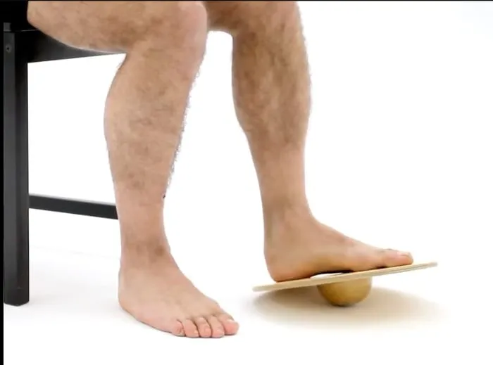 A seated person resting one bare foot on a wooden wobble board, rocking it with the ankle